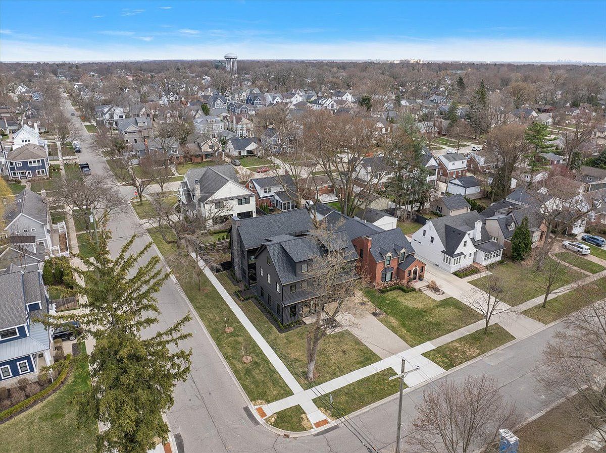 This aerial view showcases a well-maintained residential neighborhood with mature trees and diverse architectural styles. The focal point is a modern dark gray house with a contrasting red brick house next to it, both featuring manicured lawns and sidewalks. A water tower is visible in the distance, adding to the sense of community.