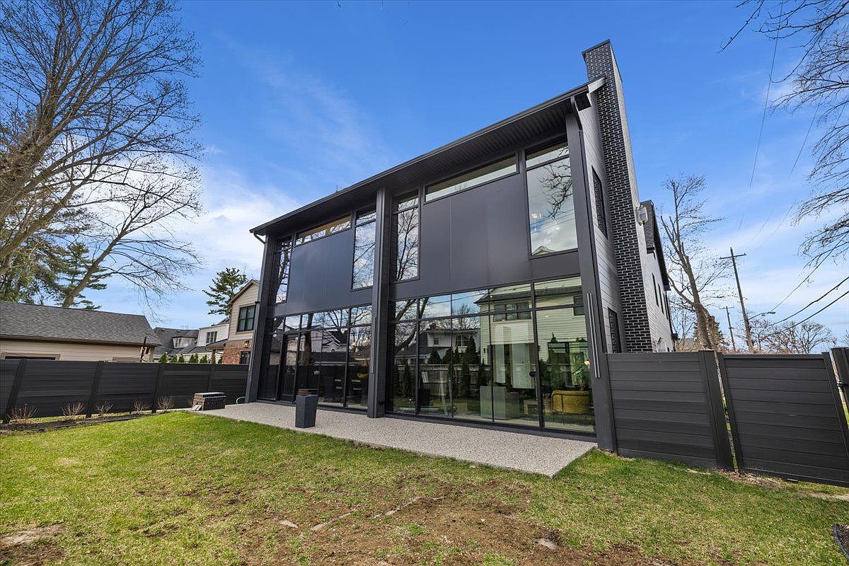 This is a rear view of a modern, two-story house with a striking black exterior and large windows. The house features a flat roof and a brick chimney. A well-maintained lawn and a dark-colored fence surround the property, adding to its contemporary appeal.