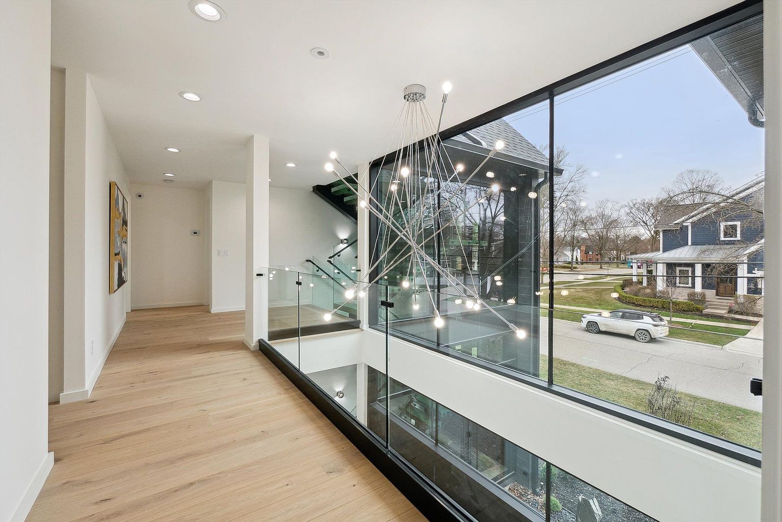 This interior shot showcases a modern hallway with light wood flooring and white walls, leading to a staircase with glass railings. A striking, multi-bulb chandelier hangs in the open space, and a large window provides a view of the neighborhood outside. The design emphasizes clean lines and natural light, creating a bright and airy atmosphere.