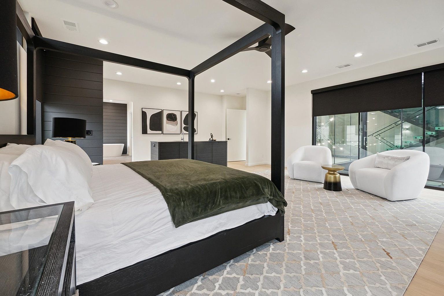 This is a primary bedroom featuring a four-poster bed with dark wood framing and an olive green throw blanket. The room includes a seating area with two white armchairs and a small side table, set upon a patterned rug. Large windows with dark shades provide natural light, and the overall design is modern and luxurious.
