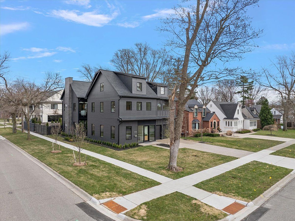 This is a front exterior view of a modern two-story house with a dark gray facade and black trim. The house features a well-manicured lawn, a concrete walkway, and mature trees in the front yard. The overall impression is one of contemporary elegance and curb appeal.