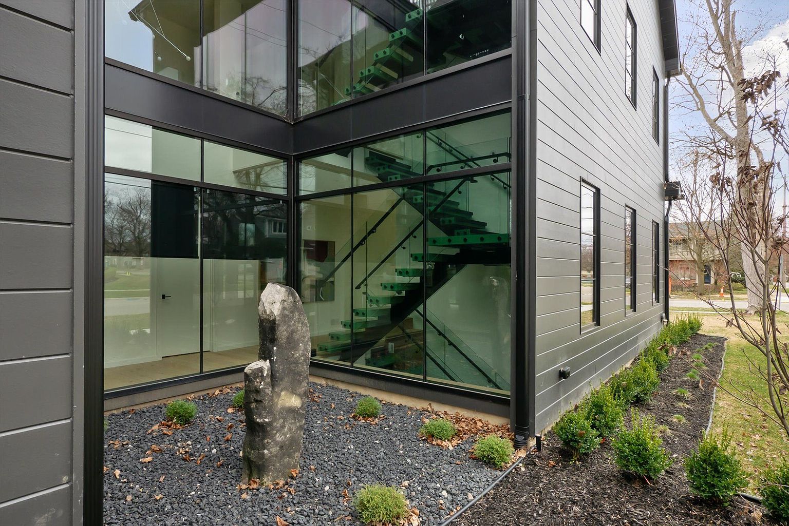 This is a side view of a modern home featuring a striking glass-enclosed staircase. The exterior is clad in gray siding, complemented by black window frames and trim. A rock garden with dark gravel and small shrubs adds a touch of natural elegance to the property's landscaping.