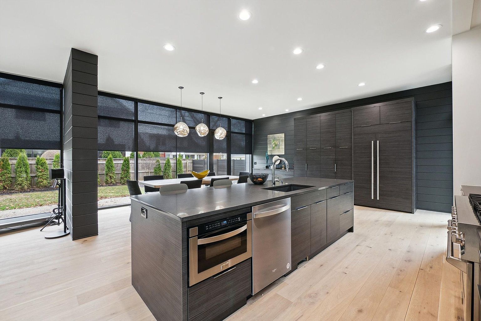 This is a modern kitchen with dark wood cabinetry and stainless steel appliances. A large island with a gray countertop dominates the center of the room, featuring a built-in microwave and dishwasher. Large windows provide ample natural light and a view of the backyard, while pendant lights hang above a dining table adjacent to the kitchen area. The perspective is a wide shot, showcasing the kitchen's layout and connection to the dining space.