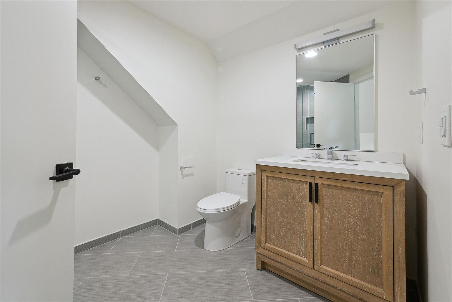 This is a well-lit guest bathroom featuring a modern vanity with a light wood finish and a white countertop, complemented by a rectangular mirror with an overhead light. A white toilet sits adjacent to the vanity, and the floor is covered in gray rectangular tiles. The walls are painted a clean white, creating a bright and inviting space.
