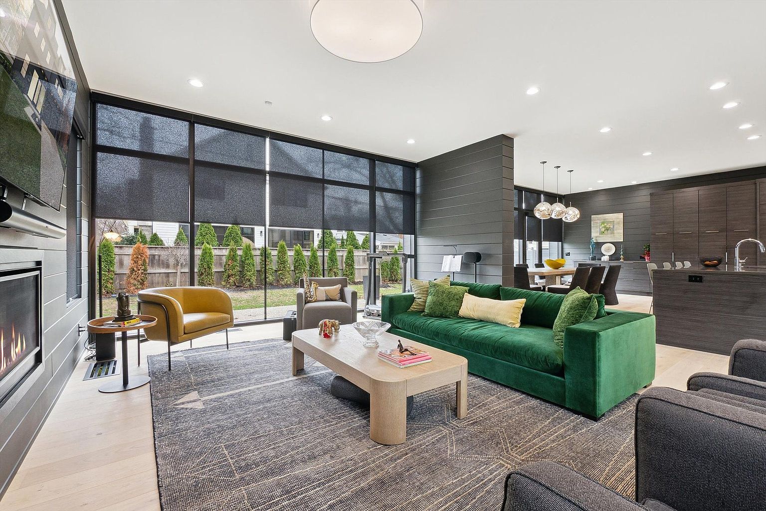 This is an interior shot of a modern living room featuring a large window with black frames and shades, offering a view of the exterior greenery. The room is furnished with a green velvet sofa, a light-colored coffee table, a yellow accent chair, and a gray patterned rug. A fireplace is visible on the left, and the open floor plan extends into the dining and kitchen areas, showcasing a cohesive and stylish living space.