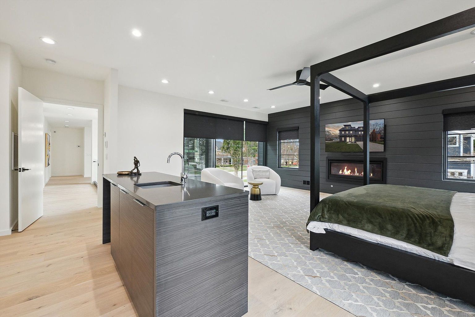 This is a primary bedroom featuring a modern design with a dark accent wall and a four-poster bed. The room includes a fireplace with a television mounted above it, and a seating area with two white chairs. A kitchen island with a sink is also present, suggesting an open-concept layout within the bedroom suite.