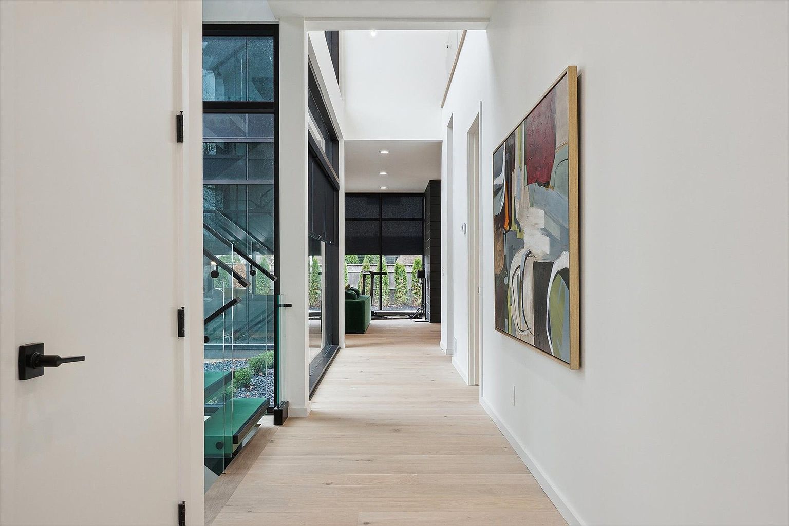 This interior shot showcases a modern hallway with light wood flooring and white walls. A striking piece of abstract art hangs on the right wall, while a glass-enclosed staircase is visible on the left, offering a view of greenery outside. The hallway leads to a bright, open space with large windows and a glimpse of a green sofa, creating a sense of depth and contemporary elegance.