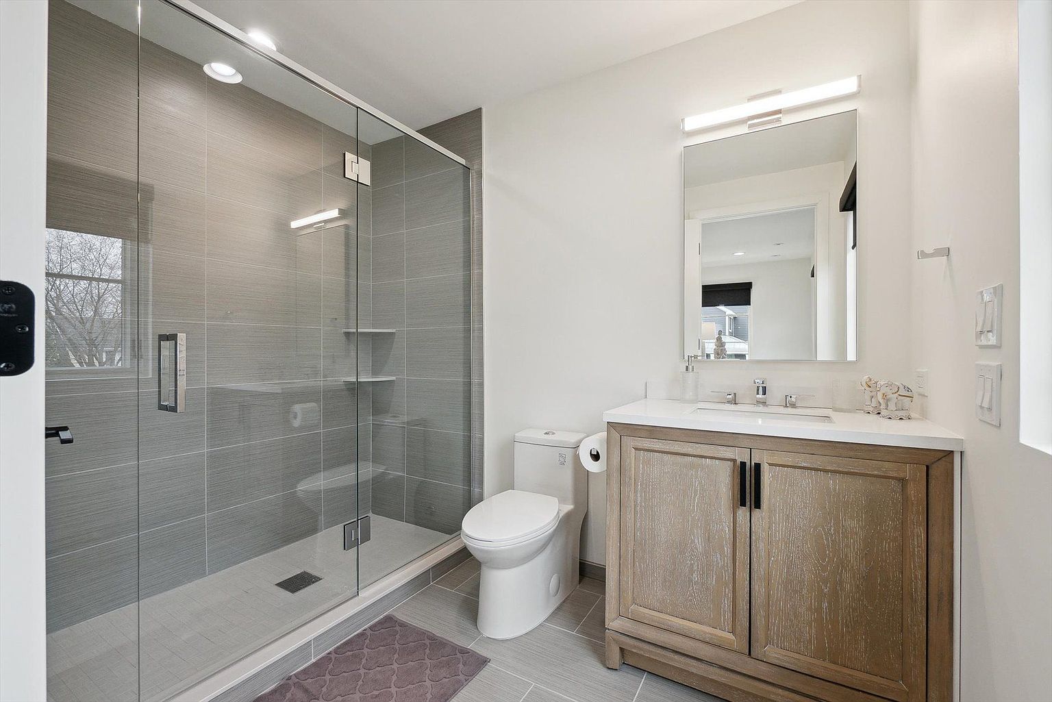 This is a well-lit bathroom featuring a glass-enclosed shower with gray tiled walls and built-in shelves. A white toilet sits next to a vanity with a light wood cabinet and a white countertop. The overall style is modern and clean, with a neutral color palette.