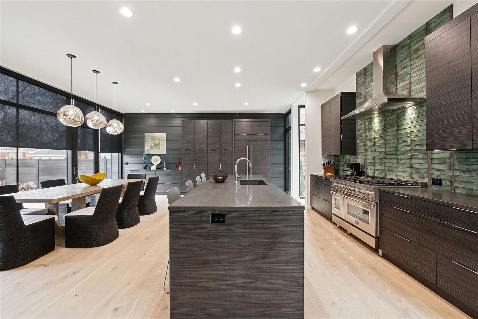 This is a modern kitchen with dark wood cabinetry and a large central island with a gray countertop. Stainless steel appliances, including a range and hood, are visible against a unique green tile backsplash. The space is well-lit with recessed lighting and pendant lights over a dining table, creating a sleek and inviting atmosphere.