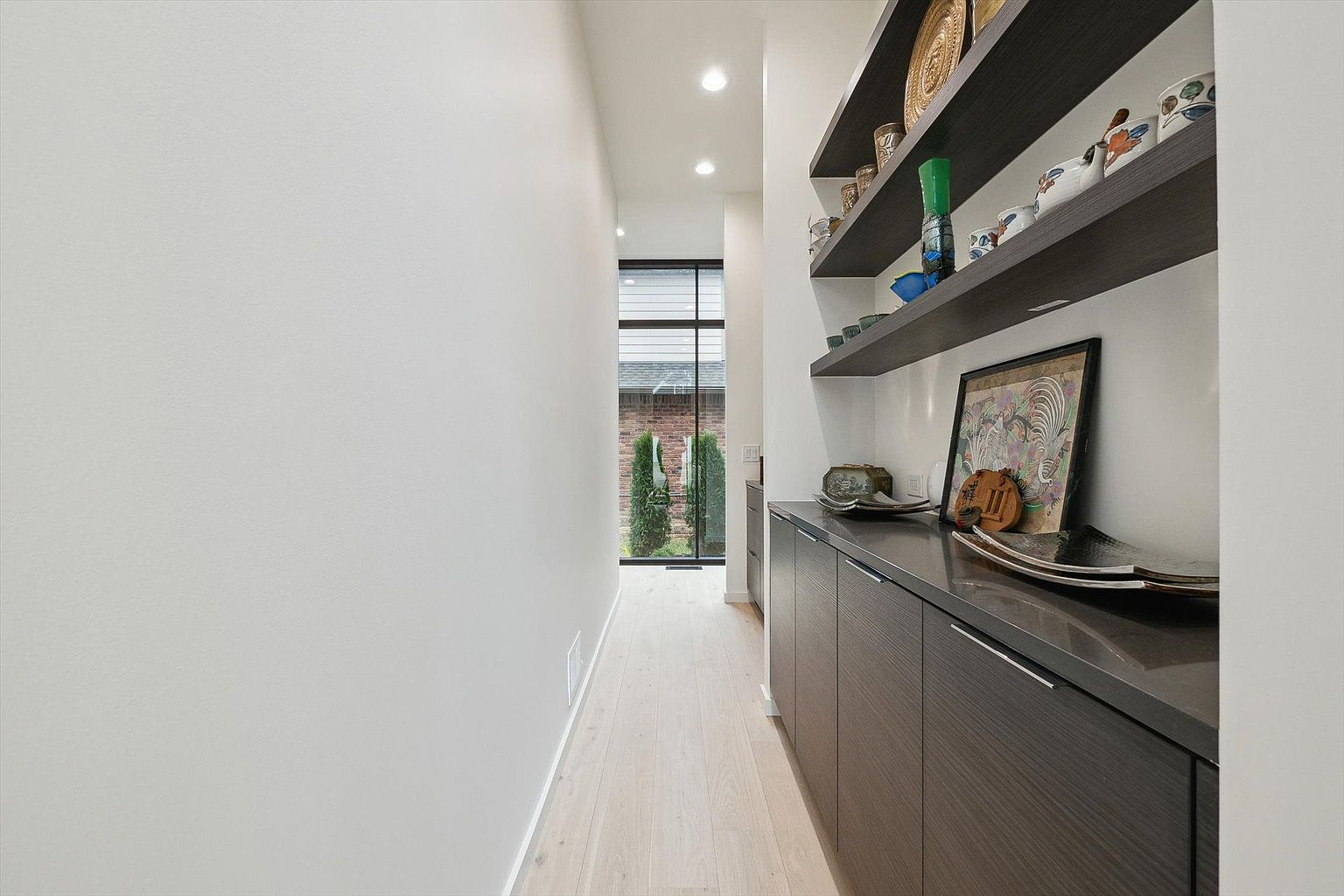 This is an interior shot of a modern hallway featuring light wood flooring and a clean, minimalist aesthetic. On the right, dark wood cabinetry with a countertop provides storage and display space, complemented by floating shelves adorned with decorative items. A window at the end of the hallway offers a glimpse of greenery, enhancing the sense of depth and natural light.