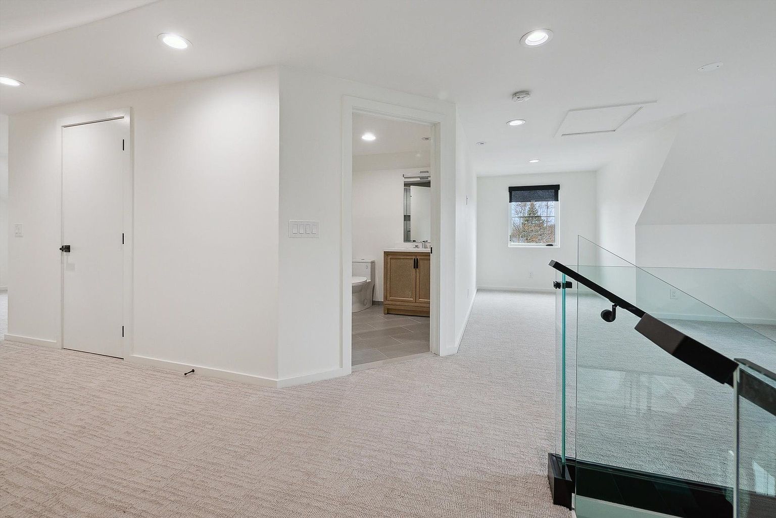 This interior shot showcases a bright hallway with neutral carpet and white walls, leading to a bathroom and another room. A glass railing with black accents provides a modern touch to the staircase. The space is well-lit with recessed lighting, creating a clean and airy atmosphere.