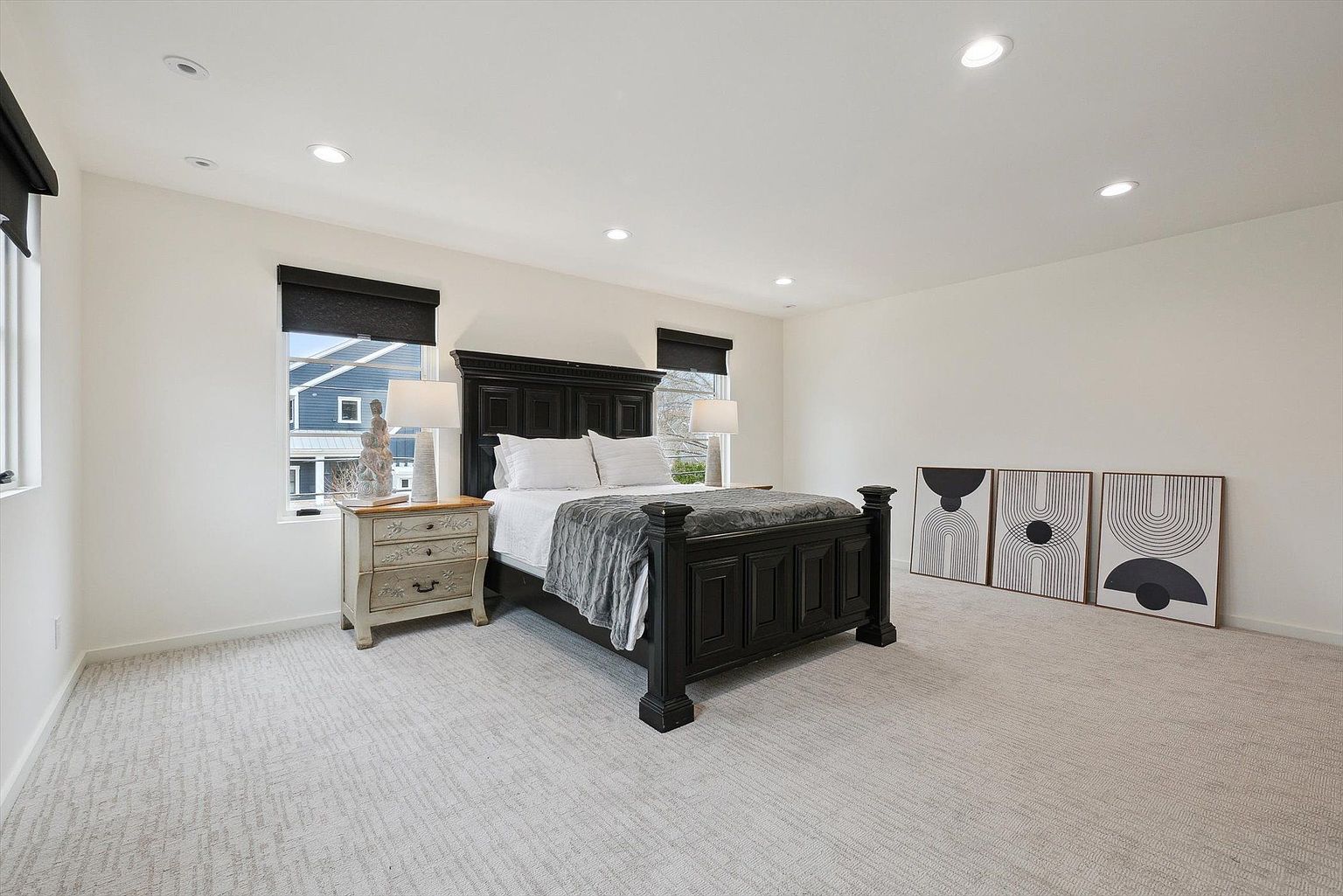 This is a spacious primary bedroom featuring a dark wood, four-poster bed with white linens and a gray throw. The room has neutral-toned carpet, white walls, and recessed lighting. Two windows with dark blinds flank a nightstand with a lamp, and three framed art pieces lean against the wall, adding a modern touch.