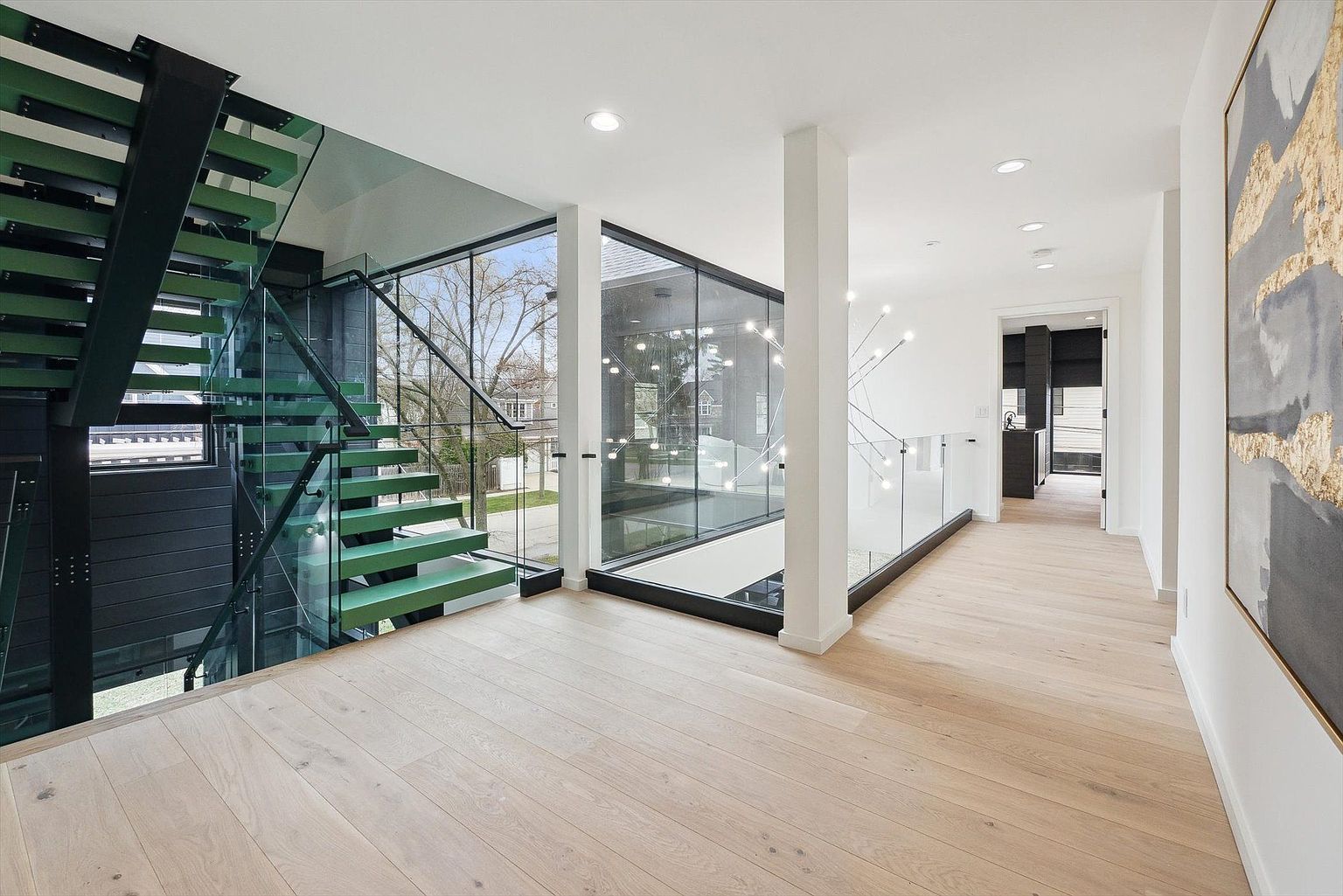 This interior shot showcases a modern hallway and staircase with a striking architectural design. The staircase features green steps and glass railings, while the hallway boasts light wood flooring, white walls, and a contemporary light fixture. Large windows provide ample natural light and views of the exterior, creating a bright and airy atmosphere.