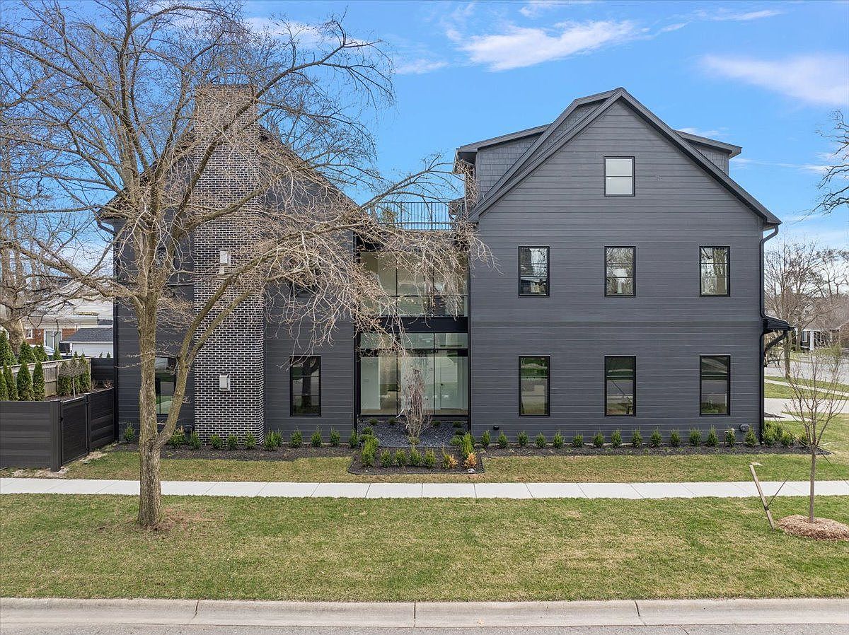 This is a front exterior view of a modern, two-story house with a dark gray facade. The house features a combination of siding and brick, with black-framed windows and a glass-enclosed entryway. A well-manicured lawn and landscaping enhance the property's curb appeal, and a mature tree stands in the front yard.