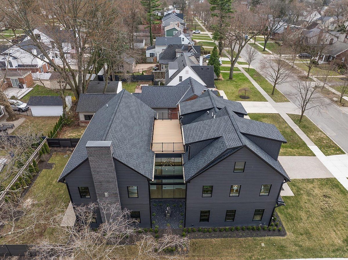 This aerial view showcases a modern, dark-colored house with a complex roofline and a rooftop deck. The property features a well-maintained lawn and landscaping, complemented by mature trees and neighboring houses. The overall impression is one of sophisticated design and comfortable suburban living.