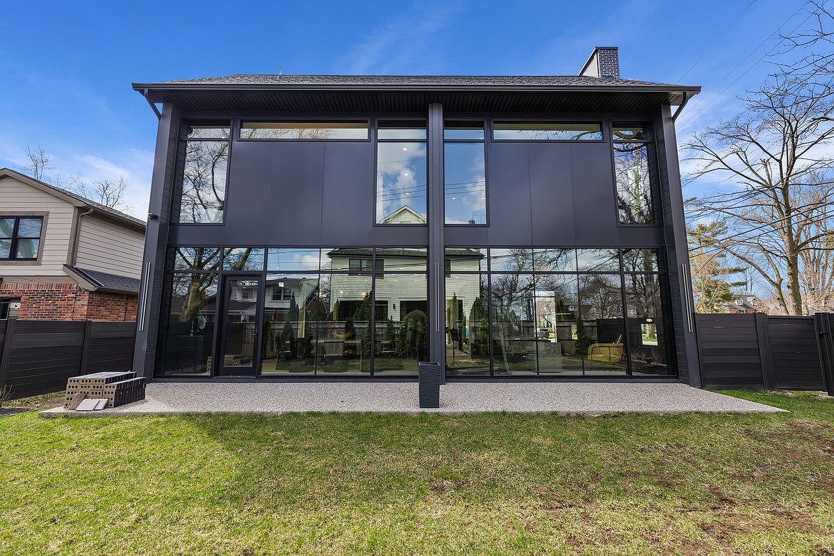 This is a rear view of a modern two-story house with a striking black exterior and expansive glass windows. The design emphasizes natural light and indoor-outdoor connectivity, with a well-maintained lawn extending from the back of the house. A dark fence encloses the property, providing privacy and security.