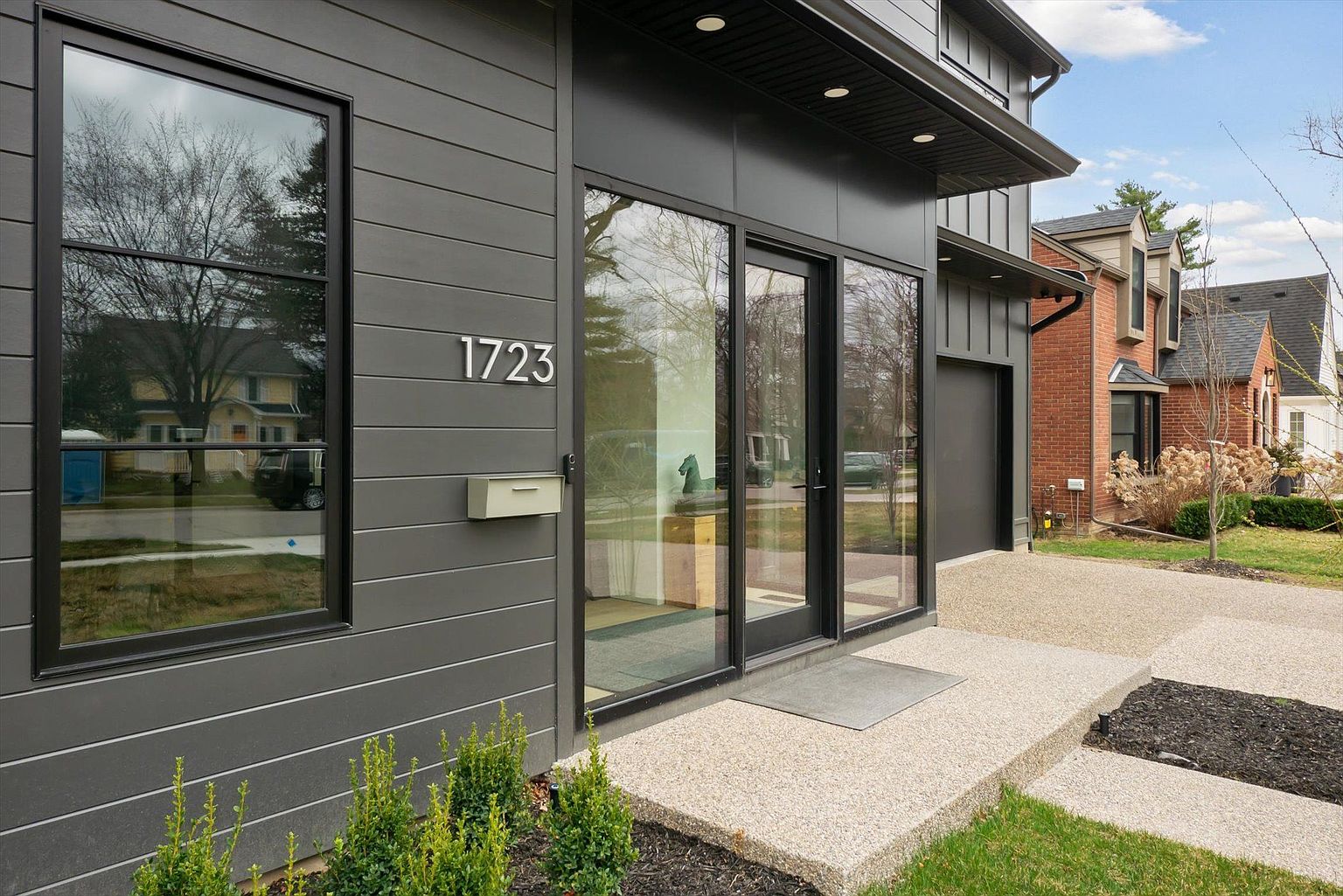 This image showcases the entryway of a modern home, featuring a sleek gray exterior with horizontal siding and black-framed windows. The house number '1723' is prominently displayed next to a minimalist mailbox. A concrete walkway leads to the glass-paneled front door, complemented by neatly trimmed greenery, creating a welcoming and stylish entrance.