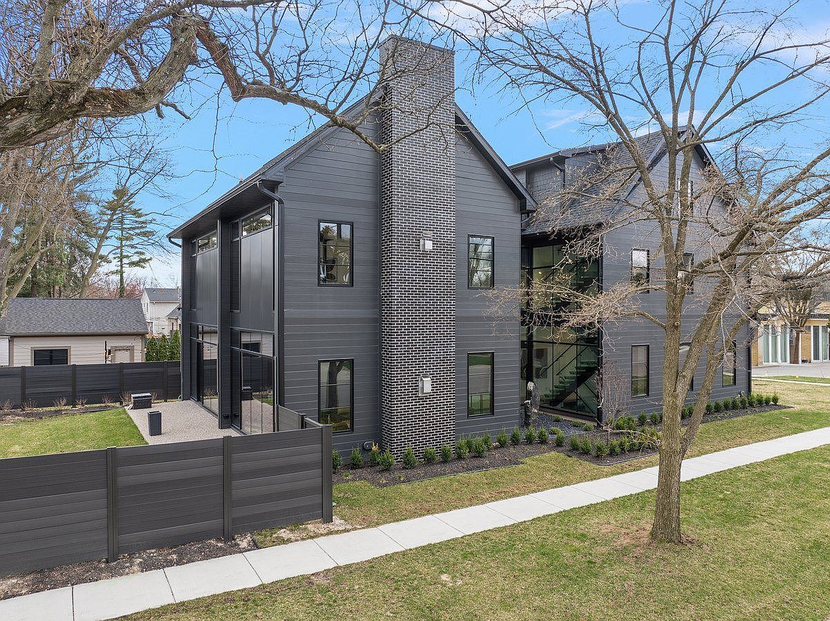 This is a front exterior view of a modern, two-story house with dark gray siding and black-framed windows. A prominent brick chimney adds a textural contrast to the facade. The property features a well-maintained lawn, a concrete walkway, and a dark gray fence, contributing to a clean and contemporary aesthetic.