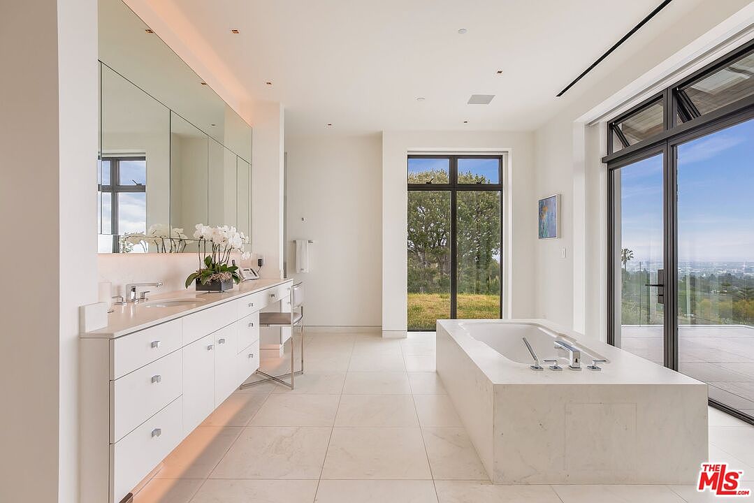 This is a luxurious primary bathroom featuring a double vanity with white cabinetry and a large mirror. A freestanding bathtub is positioned near a large window offering scenic views. The bathroom has a clean, modern aesthetic with light-colored tile flooring and ample natural light, creating a spa-like atmosphere.