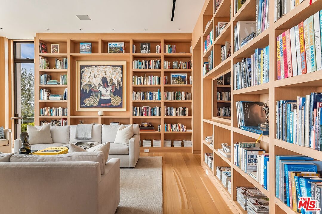 This interior shot showcases a sophisticated living room with extensive built-in bookshelves filled with books and decorative items. The room features comfortable, neutral-toned sofas and a light-colored rug, creating a cozy and inviting atmosphere. Natural light streams in, highlighting the warm wood tones of the shelving and flooring.