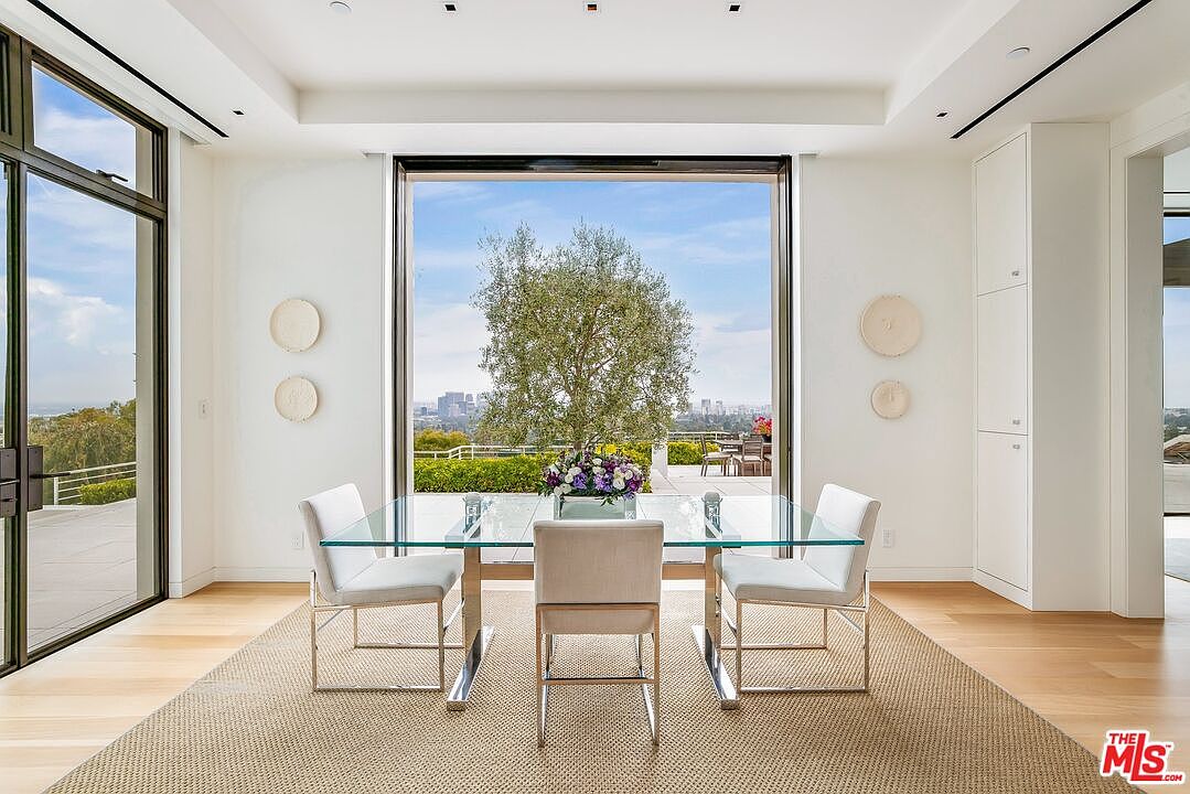 This is a bright and airy dining room featuring a glass-top table with modern chrome-legged chairs. Large sliding glass doors offer expansive views of the city skyline and a lush green landscape. The room is decorated in a minimalist style with neutral tones and natural light, creating a sophisticated and inviting atmosphere.