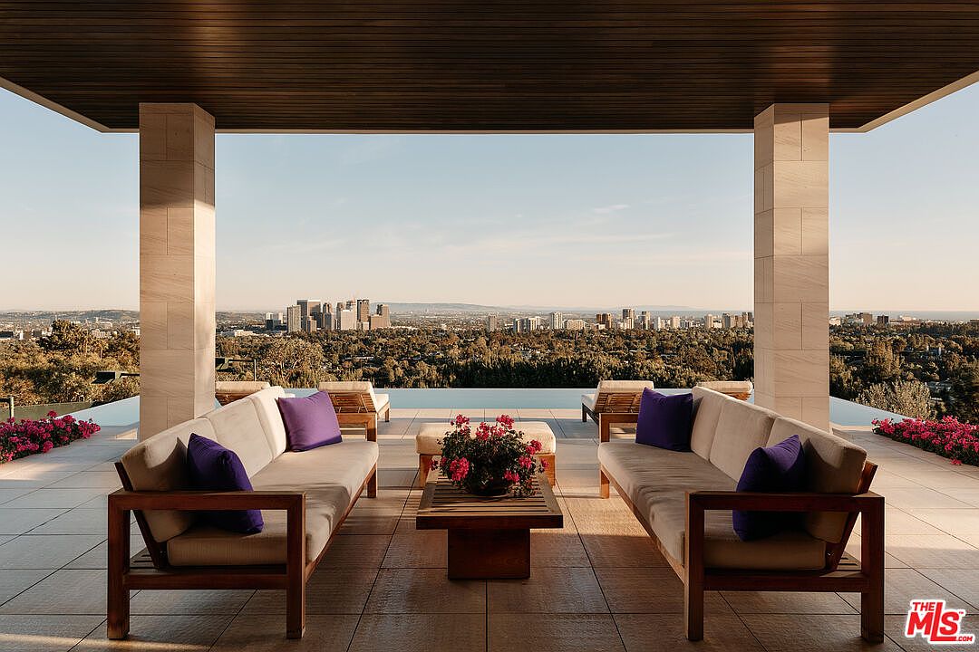 This image showcases a luxurious outdoor patio or balcony area with stunning city views. The space is furnished with two comfortable sofas adorned with purple pillows, flanking a wooden coffee table with a floral arrangement. The patio features a tiled floor and is framed by substantial stone pillars and a wooden ceiling, creating a sophisticated and inviting outdoor living space.