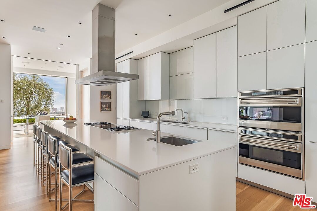 This is a bright, modern kitchen featuring sleek white cabinetry and stainless steel appliances, including double ovens and a cooktop on the large island. The island has bar seating and a stainless steel sink with a modern faucet. The kitchen has hardwood floors and a large window with a view of the city.