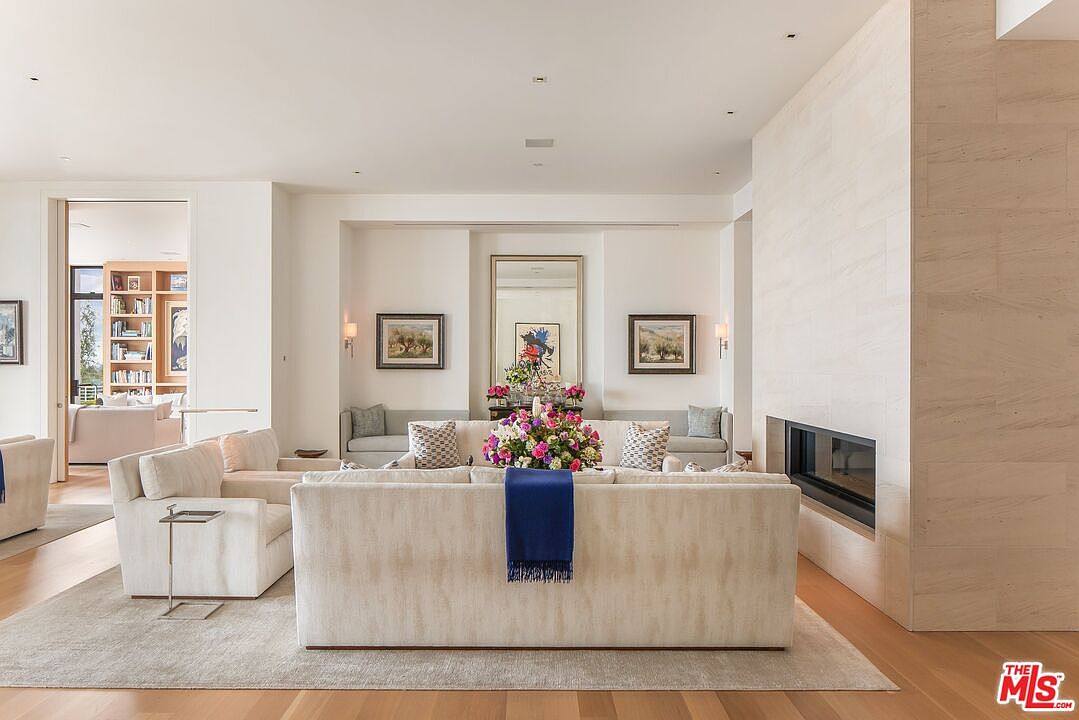 This is an interior shot of a living room featuring a neutral color palette with hardwood floors and a large area rug. The room is furnished with multiple sofas, artwork, and a modern fireplace. The space is well-lit and appears spacious, creating an inviting and elegant atmosphere.