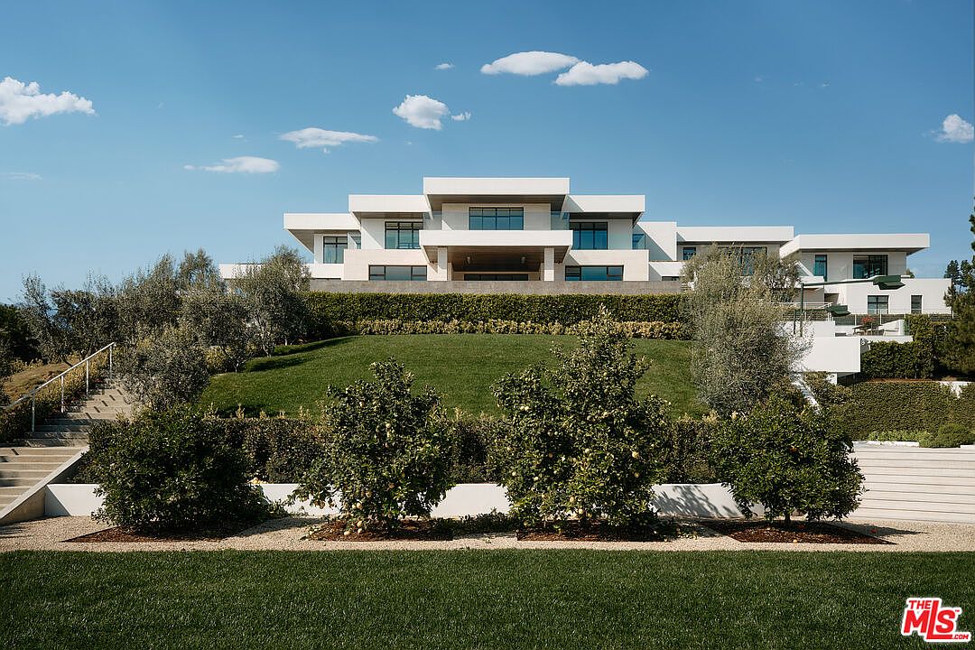 This image showcases a beautifully landscaped yard and garden area of a modern home. The property features a tiered design with lush green lawns, manicured hedges, and mature trees. A grand white modern house sits atop the hill, creating a striking focal point against the clear blue sky.