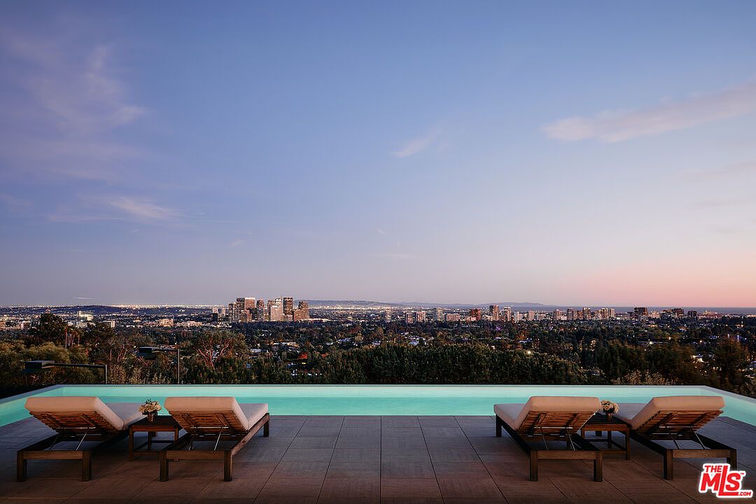This image showcases a luxurious outdoor pool area with stunning city views at dusk. The pool is surrounded by a tiled deck, furnished with elegant lounge chairs and small tables. The cityscape in the background is illuminated, creating a captivating and serene ambiance, perfect for relaxation and entertainment.