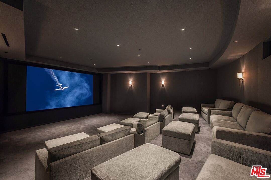 This is a home theater room featuring multiple rows of comfortable seating, including sofas and individual chairs with ottomans. A large screen displays an image of an airplane, and sconces provide ambient lighting. The room is designed for an immersive cinematic experience.