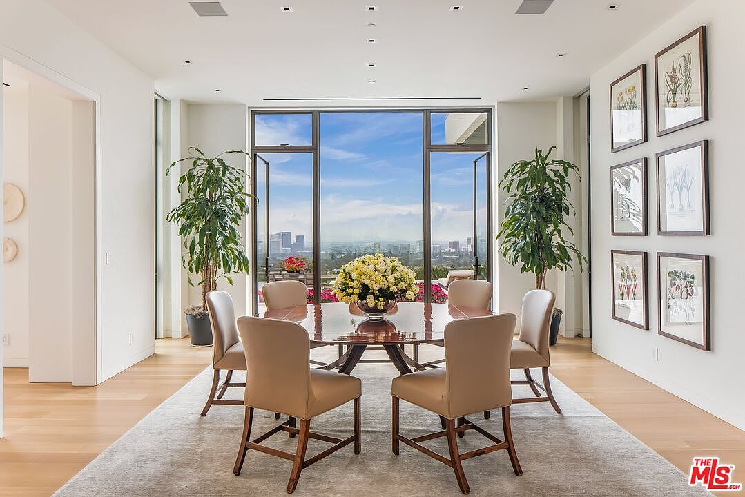 This is a bright and airy dining room featuring a round wooden table with six upholstered chairs, set on a large area rug. The room is illuminated by natural light streaming through large windows that offer a stunning city view. Two potted plants flank the windows, and framed artwork adorns the wall, adding a touch of elegance.