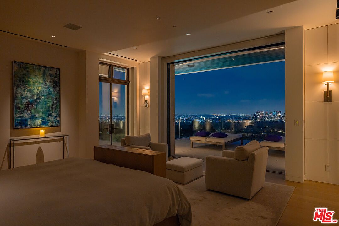 This is a luxurious primary bedroom featuring a large bed, a seating area with two armchairs and an ottoman, and a stunning view of the city skyline through a large sliding glass door leading to a balcony. The room is decorated in neutral tones, with modern lighting fixtures and an abstract painting adding visual interest. The overall impression is one of comfort, elegance, and sophistication.