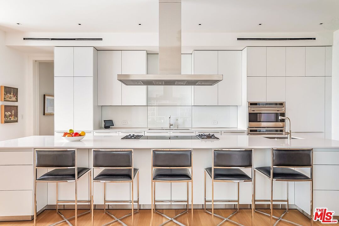 This is a bright, modern kitchen featuring sleek white cabinetry and a large island with seating for five. Stainless steel appliances and a minimalist design contribute to a clean and contemporary aesthetic. The kitchen is well-lit with recessed lighting and natural light, creating an inviting space.