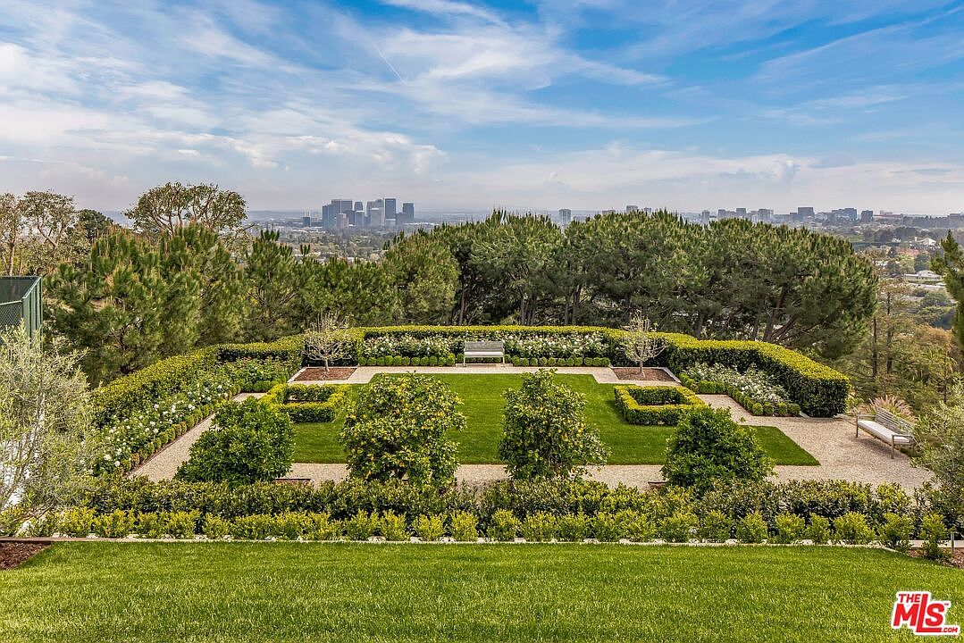 This image showcases a meticulously manicured garden with geometric hedges, gravel pathways, and mature trees, offering a serene and private outdoor space. The garden features a symmetrical design with neatly trimmed lawns and decorative plantings, complemented by a distant cityscape view. A bench provides a focal point and invites relaxation within this elegant landscape.