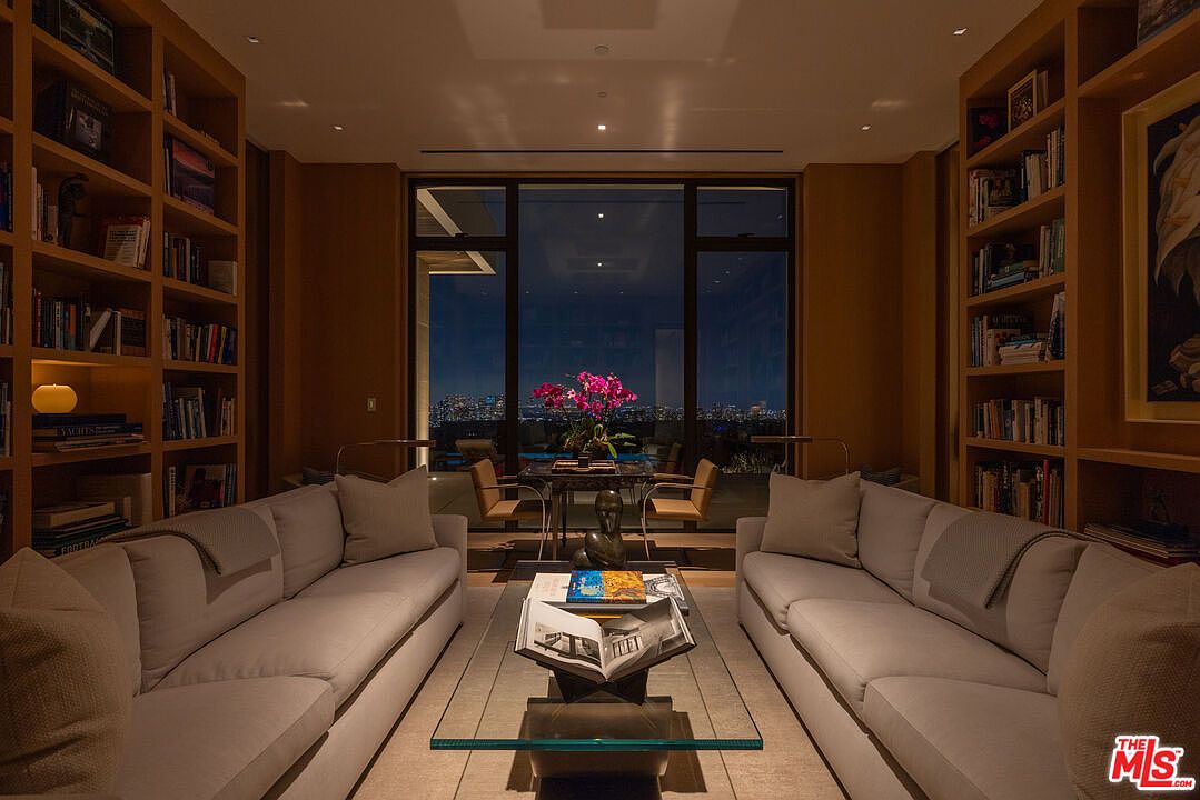 This is an interior shot of a luxurious living room featuring two light gray sofas facing each other, flanking a glass coffee table with books. The room is lined with wooden bookshelves and a large window offering a stunning night view of the city skyline. The overall impression is one of sophistication and comfort.