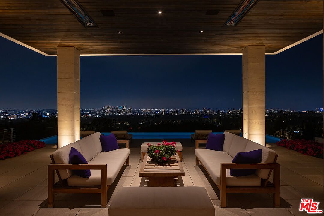 This image showcases a luxurious outdoor patio area at night, featuring comfortable seating arrangements with plush sofas and a coffee table. The patio is covered by a wooden ceiling with recessed lighting, and the open design provides stunning panoramic views of the city skyline in the distance. A pool is visible in the background, adding to the upscale ambiance.