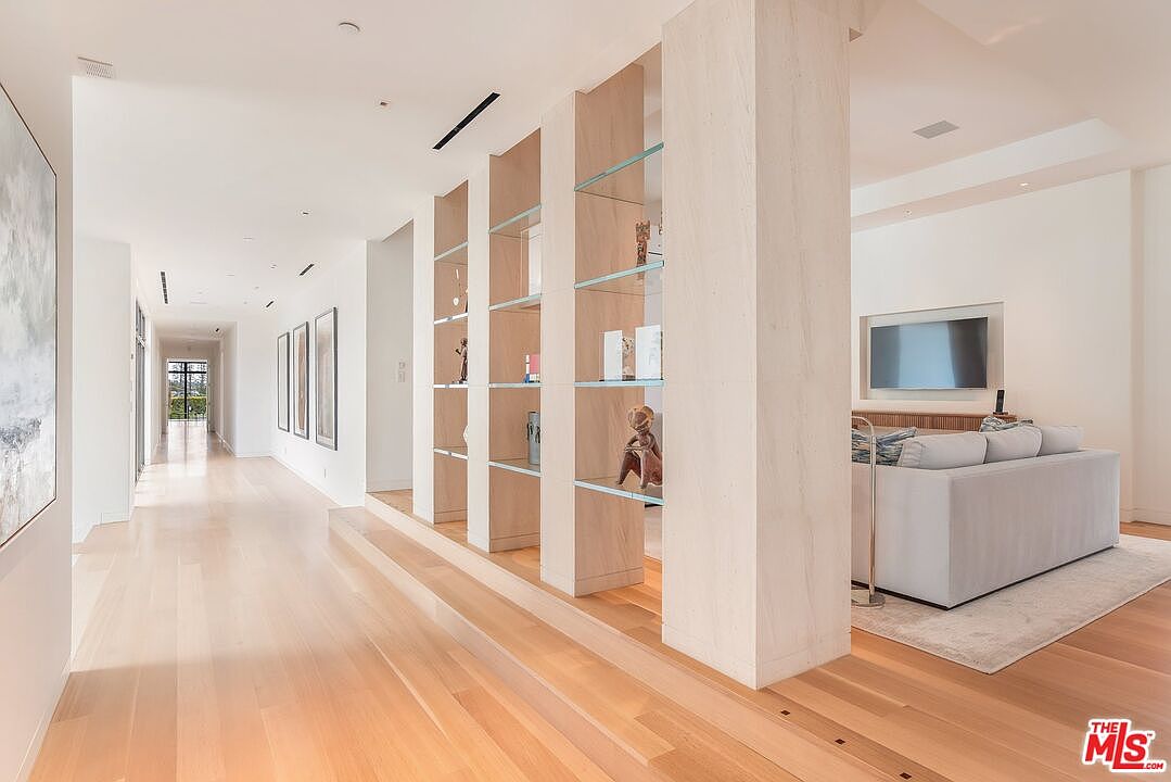 This interior shot showcases a bright and spacious hallway leading into a living area. The hallway features light wood flooring, white walls, and a unique shelving unit that partially divides the spaces. The living area includes a modern sofa and a wall-mounted television, creating a seamless transition between the two areas.