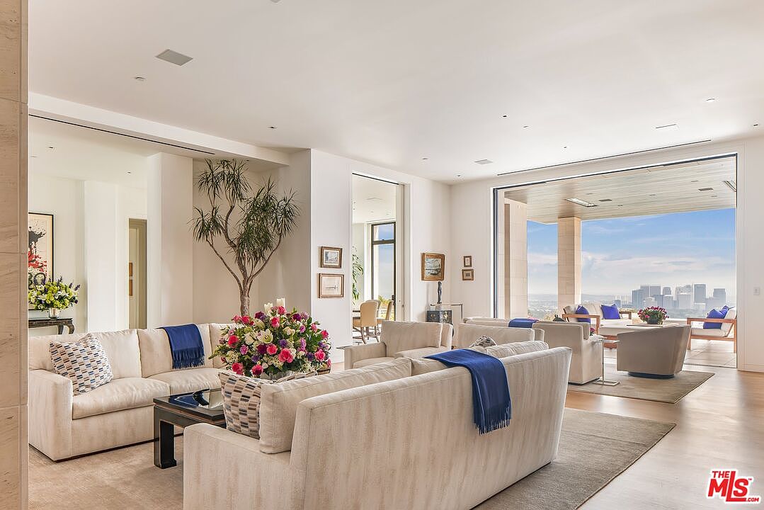 This is a bright and spacious living room featuring neutral-toned sofas, a dark wood coffee table, and a large floral arrangement. The room opens to an outdoor balcony with city views, creating an indoor-outdoor living experience. The style is modern and luxurious, emphasizing natural light and open space.