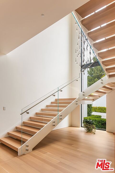 This interior shot showcases a modern staircase with wooden steps and a glass railing, creating an open and airy feel. Natural light floods the space through a large window, highlighting the clean lines and minimalist design. The hardwood flooring complements the warm tones of the steps, adding to the inviting atmosphere.