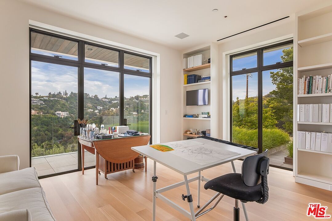 This is a bright and airy office space featuring large windows with views of the surrounding landscape. The room includes a drafting table, a drop-leaf table with art supplies, built-in shelving, and a comfortable chair. The light wood flooring and neutral color palette create a calm and productive atmosphere.