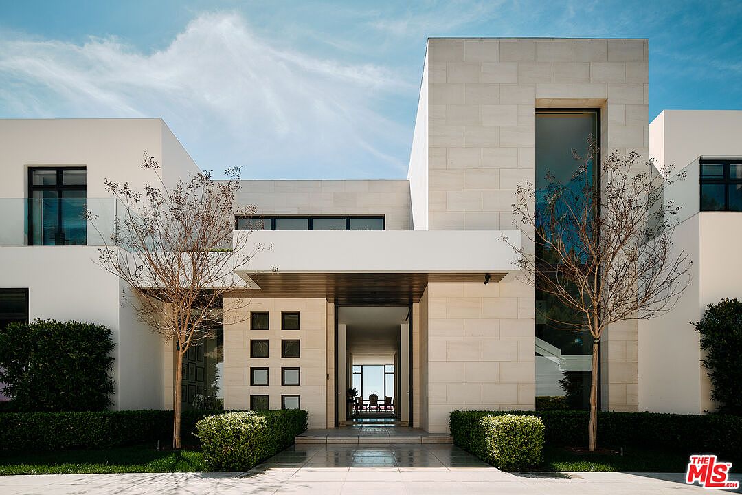 This image showcases the grand entryway of a modern home, featuring a symmetrical design with clean lines and a neutral color palette. The entrance is framed by manicured hedges and trees, leading to a covered walkway with a series of square windows on one side. The open doorway reveals a glimpse of the interior, suggesting a spacious and well-lit living area.