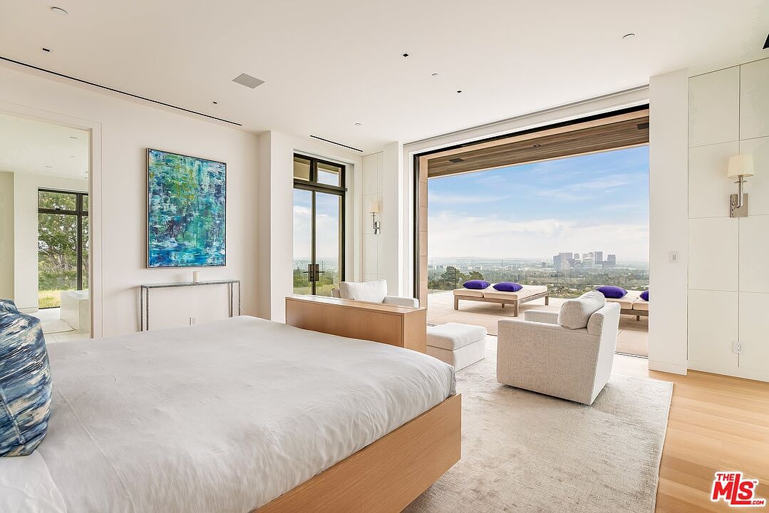 This is a primary bedroom featuring a large bed with white linens, a modern abstract painting, and a seating area with a light-colored armchair and ottoman. A sliding glass door opens to a balcony with lounge chairs and a city view. The room has a minimalist design with light wood floors and white walls, creating a bright and airy atmosphere.