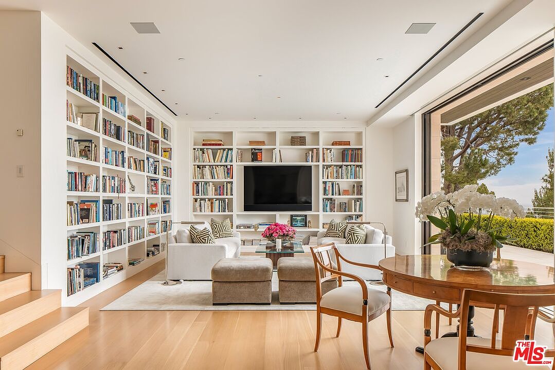 This is a bright and spacious living room featuring floor-to-ceiling bookshelves filled with books, flanking a large flat-screen television. Two white sofas face each other across a light-colored rug, with ottoman-style seating in between. A round wooden table with chairs sits in the foreground, adding a touch of elegance to the room.