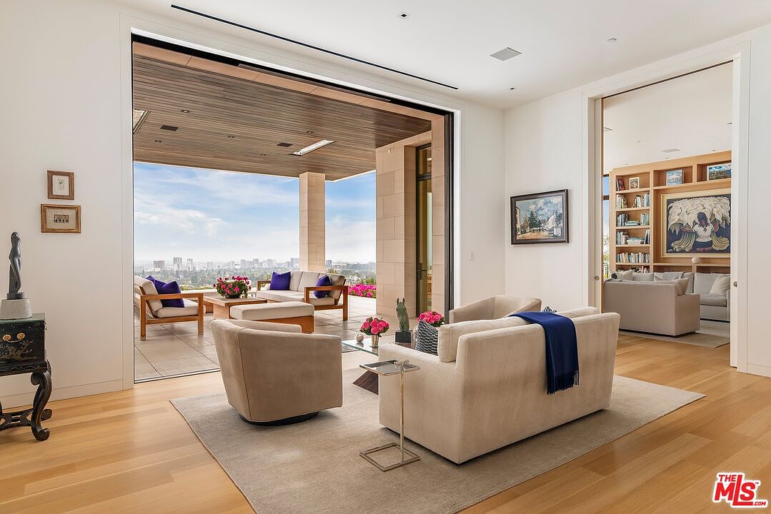 This is a luxurious living room that seamlessly blends indoor and outdoor spaces with a large sliding glass door leading to a patio with city views. The interior features modern furniture, including a beige sofa and swivel chairs, complemented by a neutral-toned rug and hardwood flooring. The open layout connects to another living space, enhancing the sense of spaciousness and elegance.