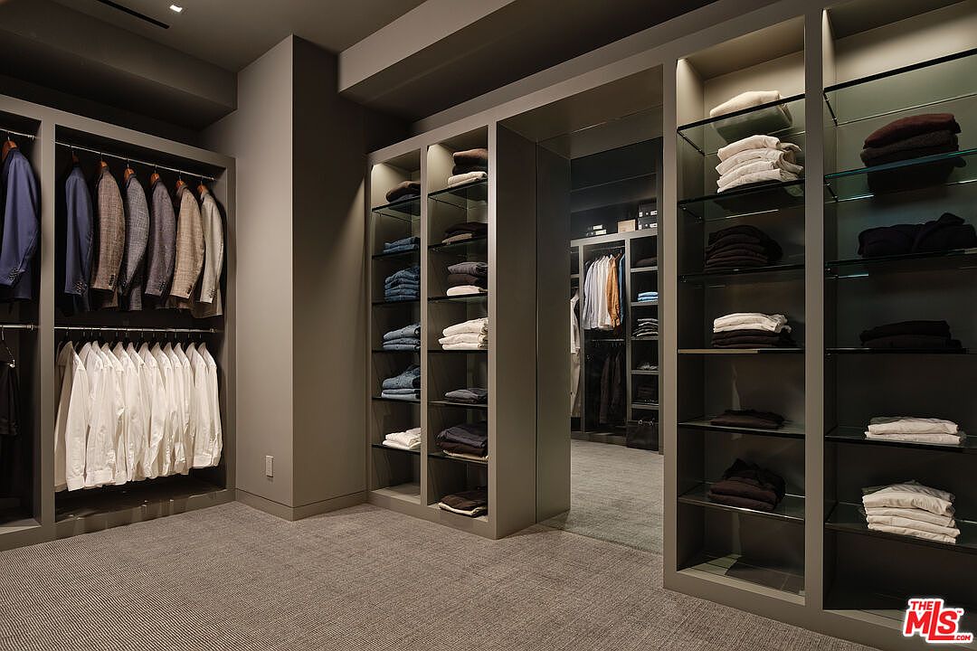 This is a well-organized walk-in closet featuring custom shelving and hanging rods. The closet is filled with neatly arranged clothing, including suits, shirts, and folded items. The neutral color palette and ample lighting create a sophisticated and functional space.