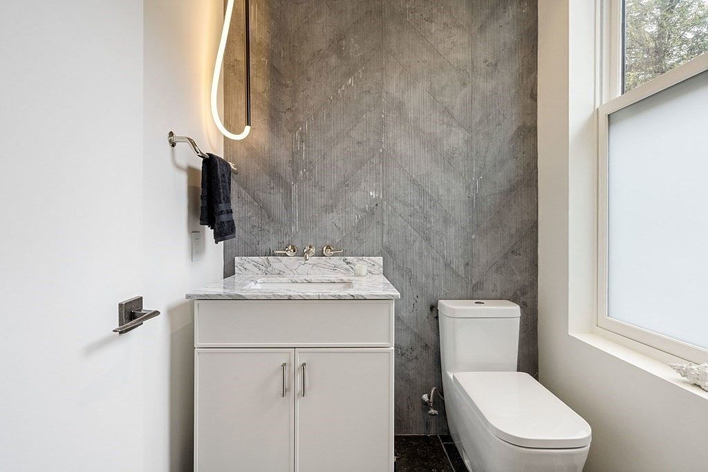 This is a modern guest bathroom featuring a white vanity with a marble countertop and a vessel sink. The wall behind the vanity is covered in gray chevron-patterned tiles, adding a touch of sophistication. A sleek, minimalist toilet sits beside a window, which provides natural light, and a unique, curved light fixture illuminates the space.