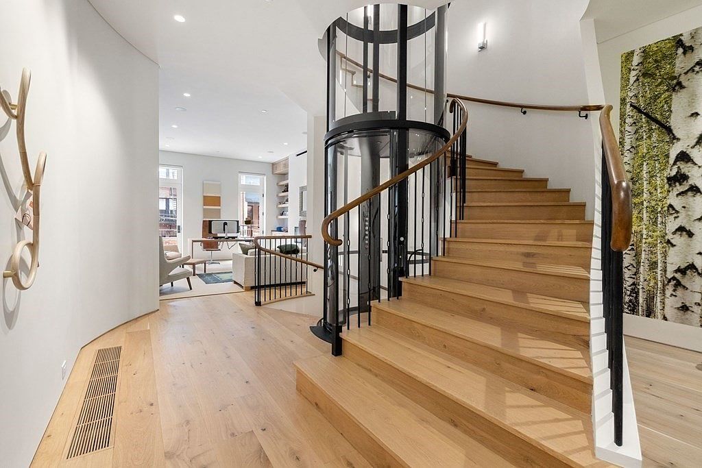 This interior shot showcases a modern home's hallway and staircase, featuring light wood flooring and a unique cylindrical elevator. The staircase has wooden steps and a black railing with a wooden handrail, while the elevator is encased in glass and black metal. The space is bright and airy, leading into a living area visible in the background.