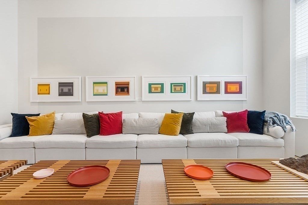 The image showcases a well-lit living room featuring a large, modular white sofa adorned with an array of colorful pillows. Above the sofa, four framed artworks add a touch of modern art. Two wooden slat coffee tables with red decorative plates sit in front of the sofa, completing the inviting and stylish living space.