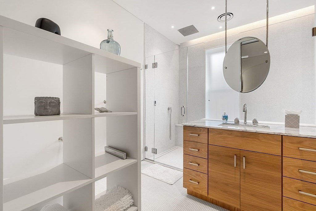 This is a bright and modern primary bathroom featuring a glass-enclosed shower, a floating wood vanity with a marble countertop, and a round mirror suspended from the ceiling. The walls and floor are covered in small white tiles, creating a clean and minimalist aesthetic. A white shelving unit provides storage and display space, adding to the room's functionality and style.
